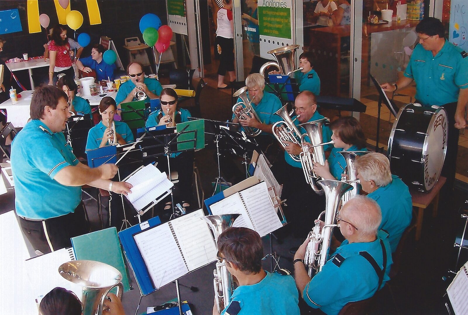 Beenleigh Brass Band in teal uniforms performing at a community event, conductor in the foreground